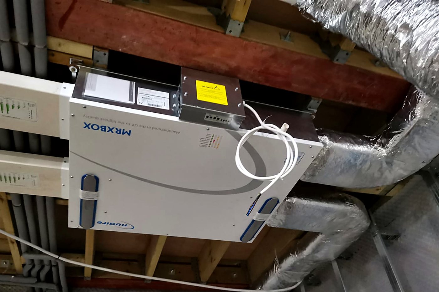 A Nuaire MRXBOX heat recovery unit suspended in a ceiling void between timber joists, with insulated ducting run over the top of it